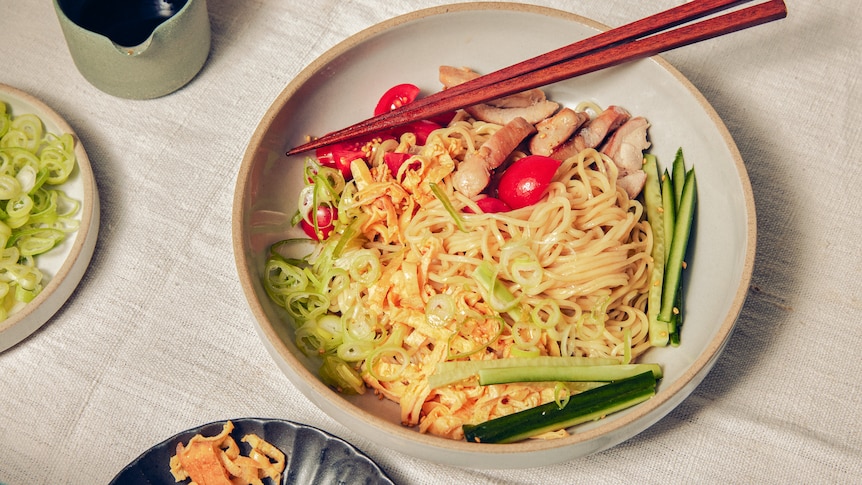 Chopsticks rest on a serving bowl of noodles topped with chicken, tomatoes and cucumber.