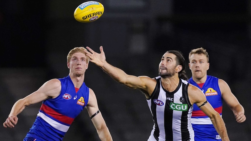 An AFL ruckman gets his hand to the ball to tap it clear while his opponent watches.