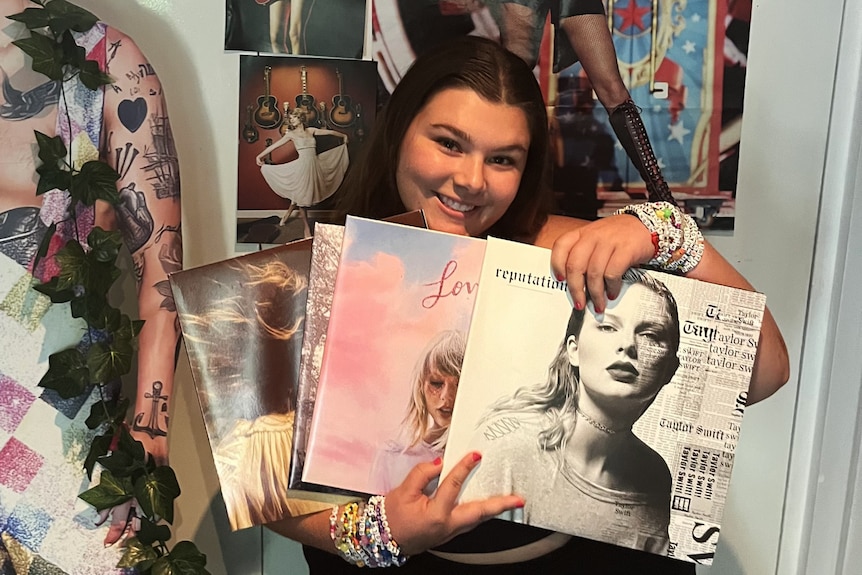 A woman smiles and holds up four Taylor Swift records in front of her.