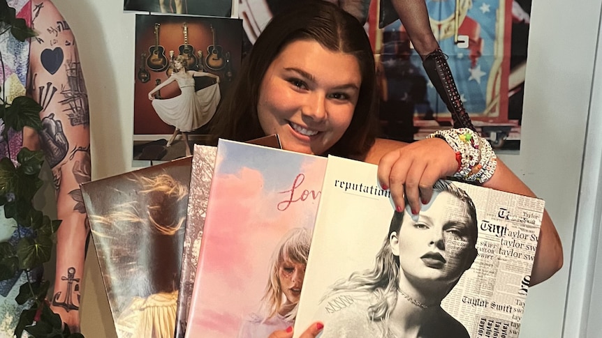 A woman smiles and holds up four Taylor Swift records in front of her.