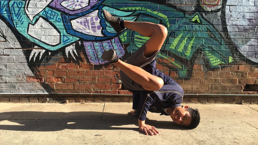 Photograph of 2019 Heywire winner Ivan Reyes break dancing in front or a wall with a mural.