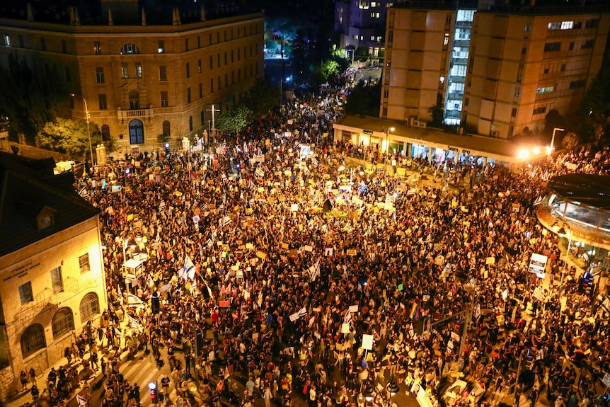 An aerial shot shows thousand of people gathered closely in a town square under lights at night.