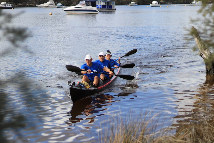 A long shot of three people in blue t-shirts kayaking in a river, boats, greenery in the background, 