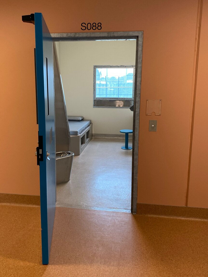 An open door to a prison cell, with a bed, toilet and seat inside.