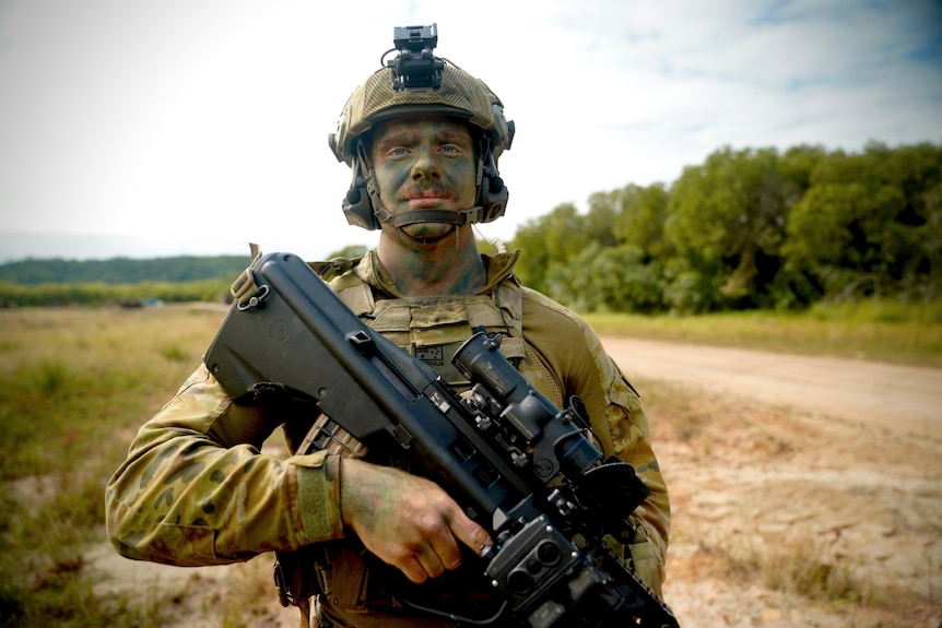 An infantry soldier in battle dress and face camouflage looks at the camera as he holds his weapon while on patrol. 