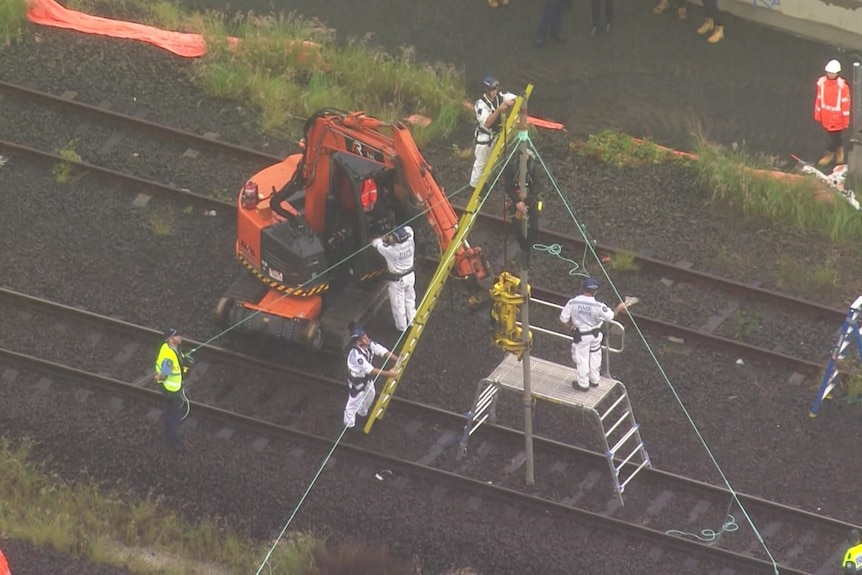 A man at the top of a pole over a train line with machinery and police below