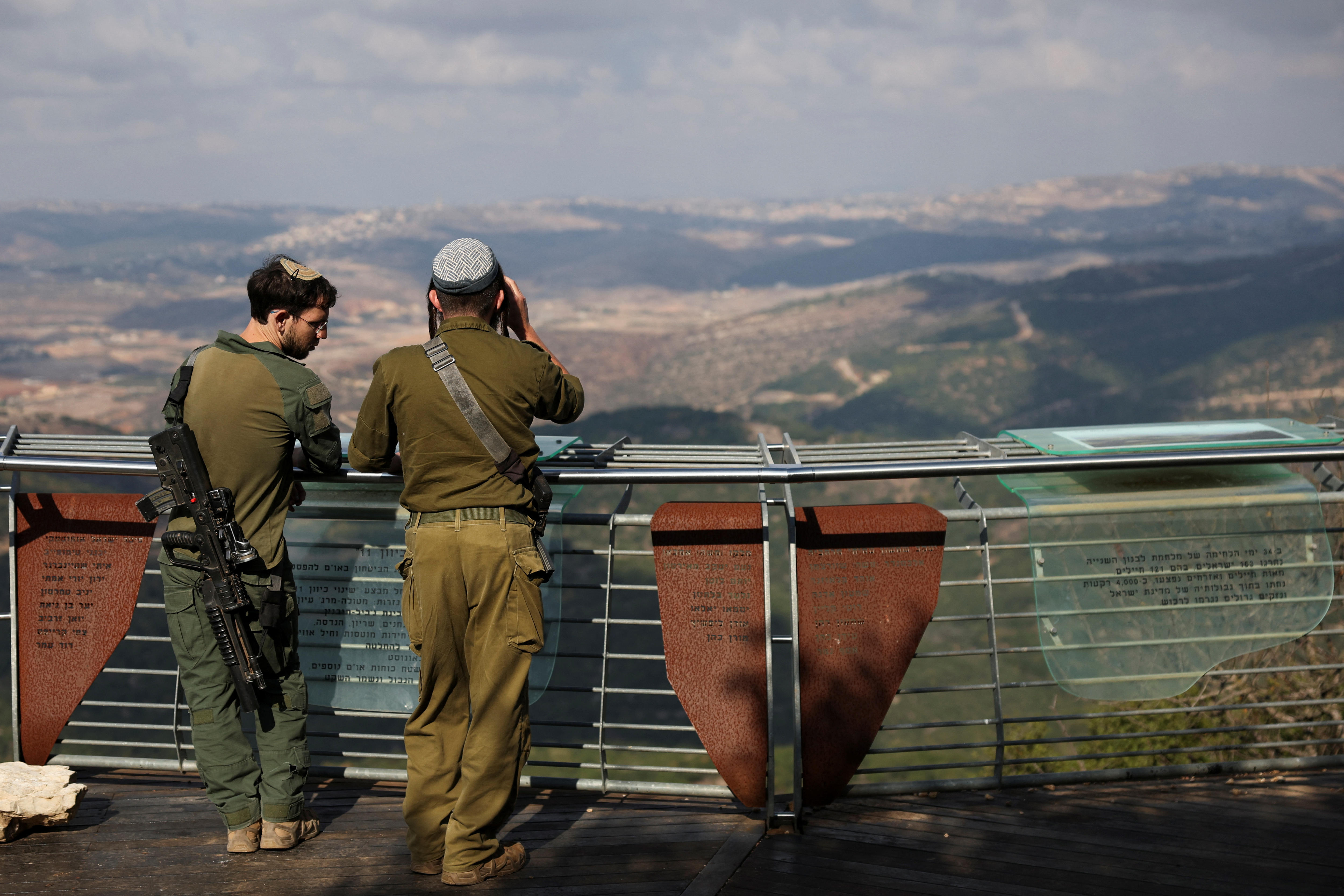 Two Israeli soldiers standing on a look out with binoculars, backs turned to the camera, looking over a vista.