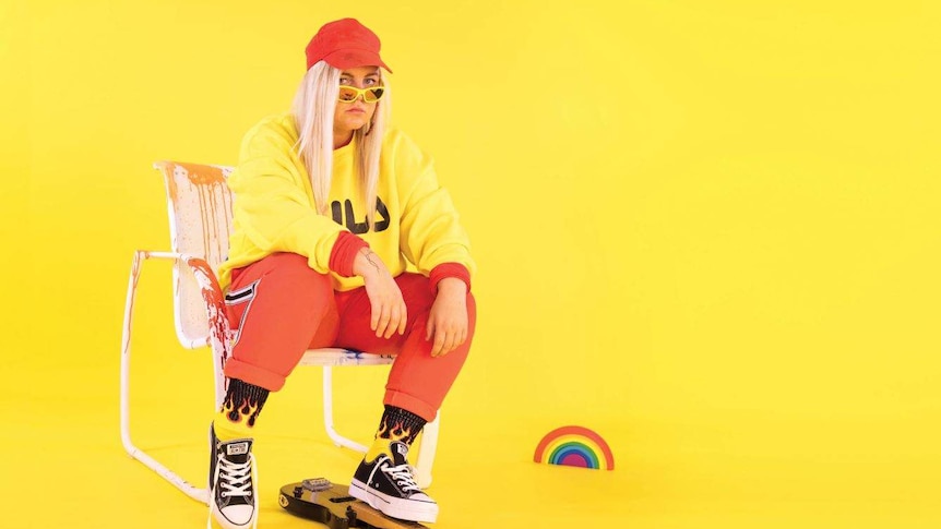 A woman with blonde hair and an orange cap sigs on a chair in a yellow room
