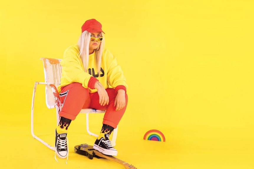 A woman with blonde hair and an orange cap sigs on a chair in a yellow room