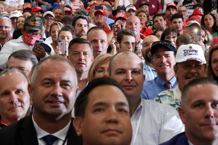 A large crowd of Donald Trump supporters gather to watch a rally. Most are smiling, some are holding up their phones to film