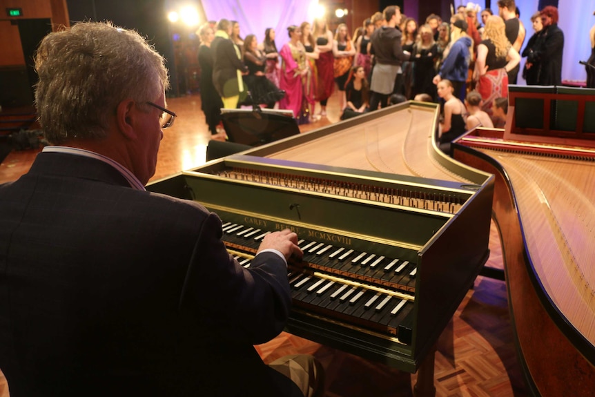 A man plays the piano with opera cast members in the background.