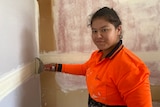 A smiling young woman wearing a neon orange t-shirt plasters a wall with a tool, looks at the camera.