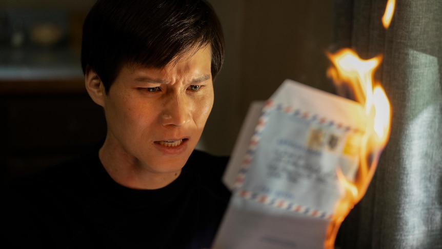 A film still of Hoa Xuande, a Vietnamese Australian man with a distressed expression, holding a burning letter.