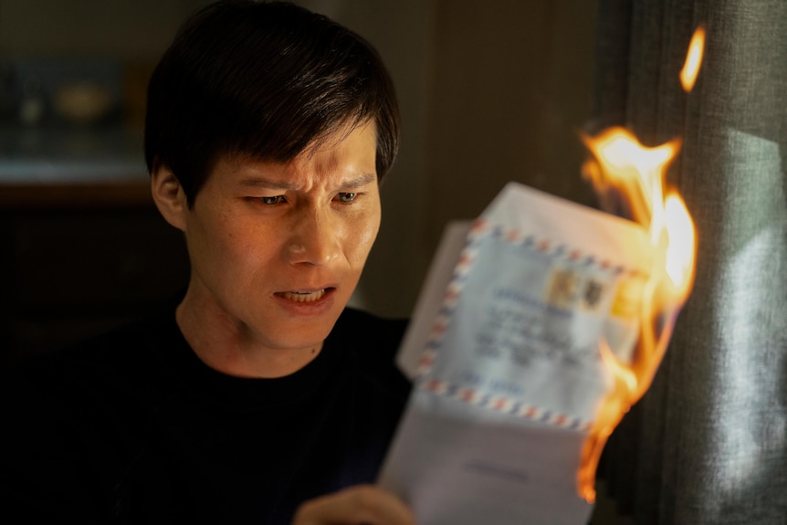 A film still of Hoa Xuande, a Vietnamese Australian man with a distressed expression, holding a burning letter.