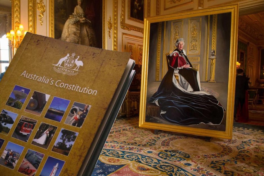 A book that says "Australia's Constitution" creeps into frame of a wide shot showing a large portrait of the Queen.