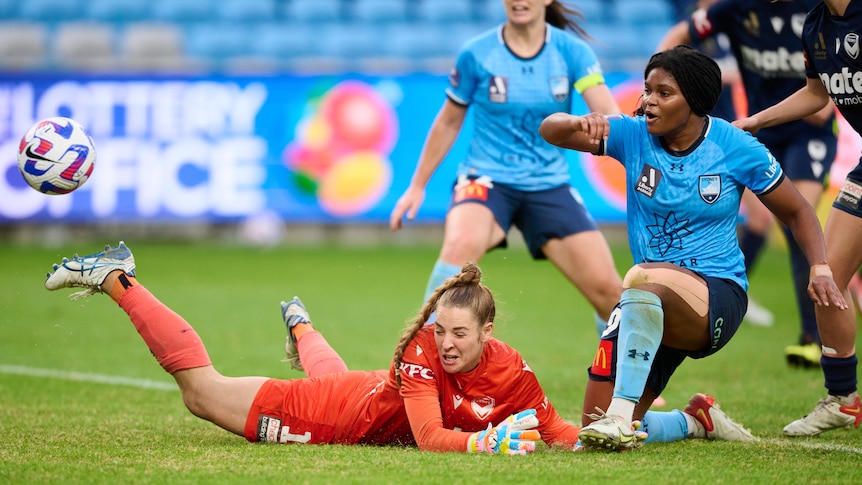 A soccer player wearing light blue kicks the ball past a goalkeeper wearing bright orange during a game