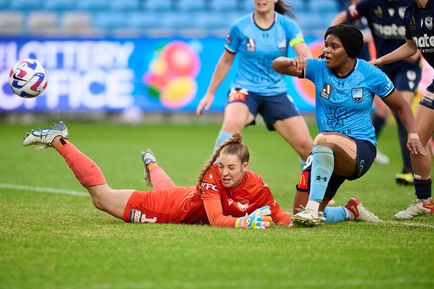 A soccer player wearing light blue kicks the ball past a goalkeeper wearing bright orange during a game