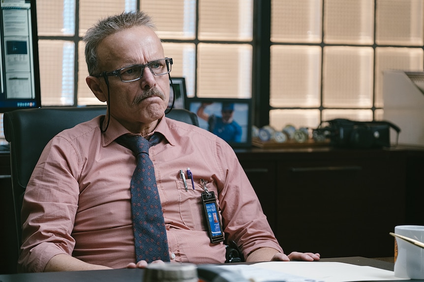 A bespectacled man in shirt and tie with greying moustache, short hair and stern expression sits in at desk in office.