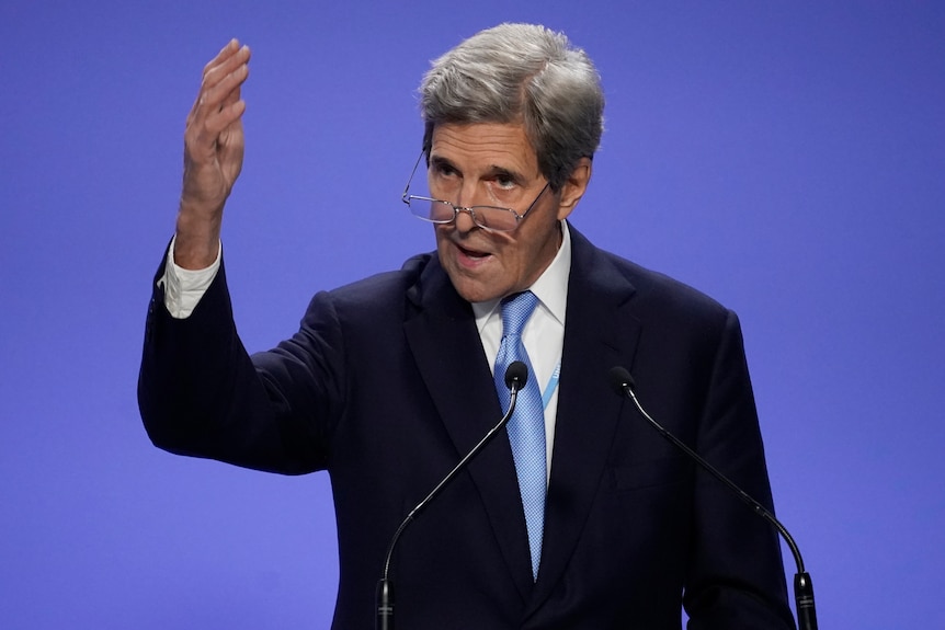 John Kerry stands at a lectern at COP26, gesturing with his hand