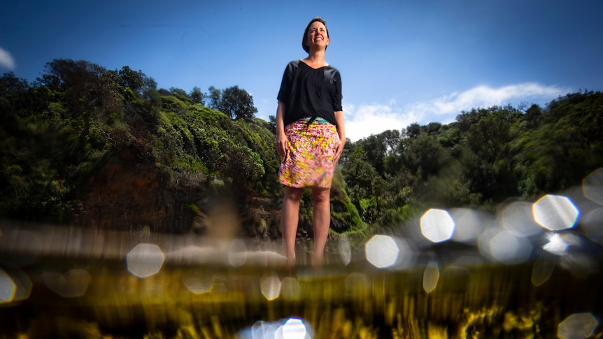 Michelle Voyer stands in a patterned skirt and black top by the water's edge with the tide out of focus in the foreground.