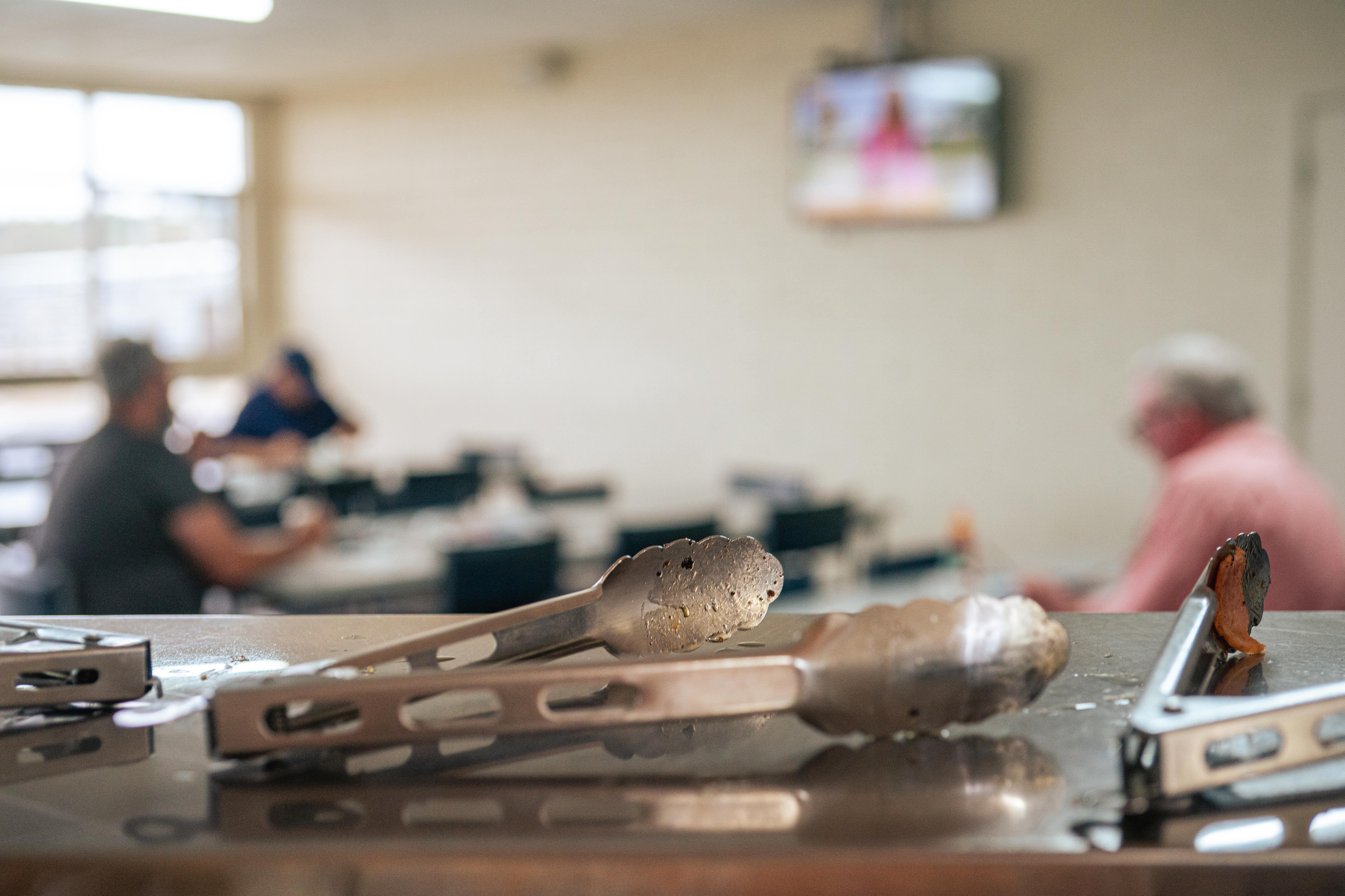 Pinzas de cocina sobre un banco y, al fondo, visitantes en los almacenes. 