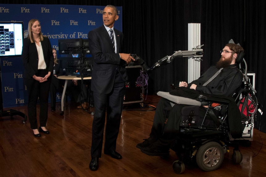 Barack Obama shakes hands with a robotic arm controlled by Nathan Copeland who is sitting in a wheelchair.