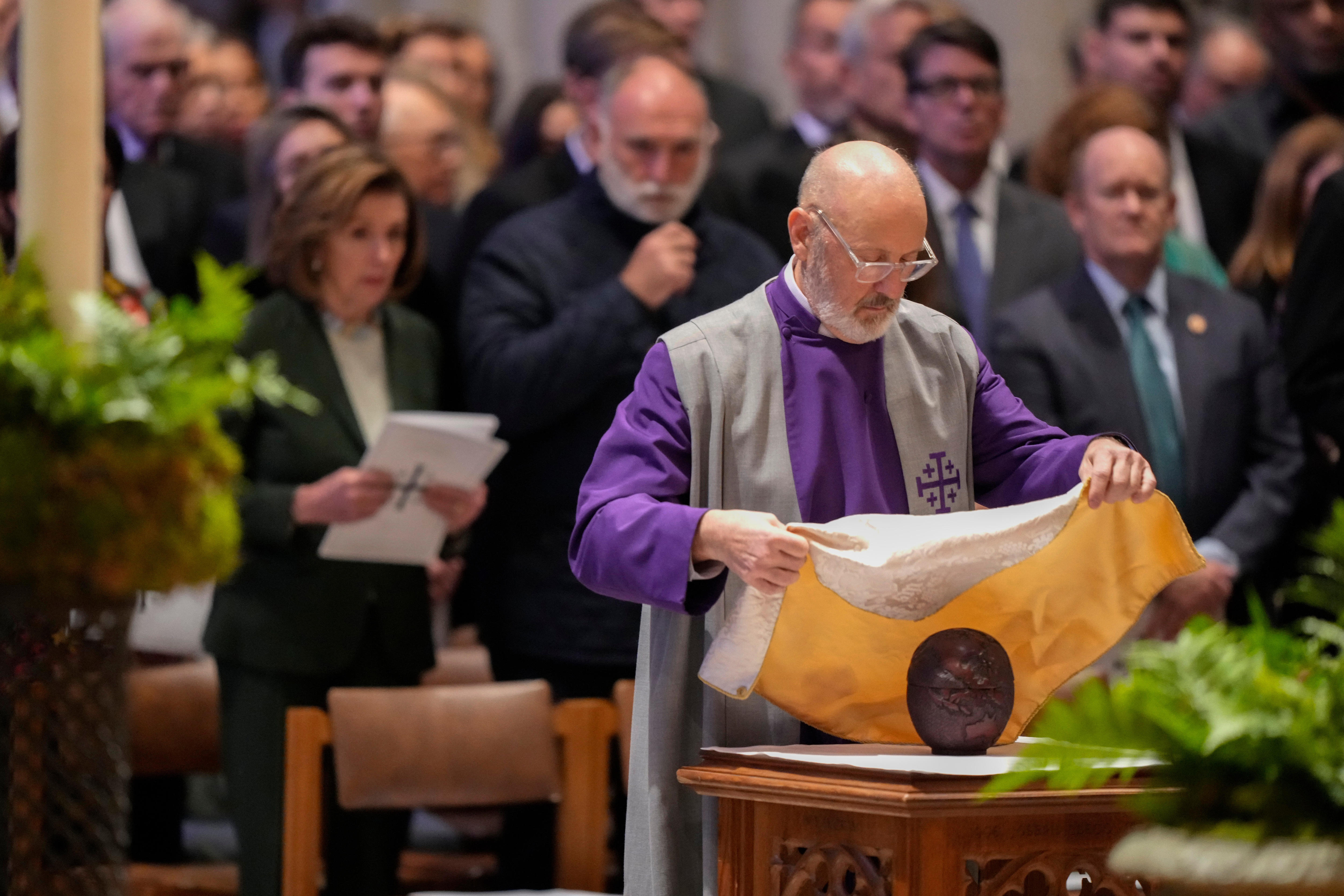 A male priest in ceremonial robes lays a piece of cloth over an urn at an altar in a cathedral while a crowd watches.