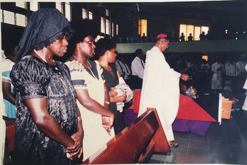 An old photo of women watching a priest next to a coffin