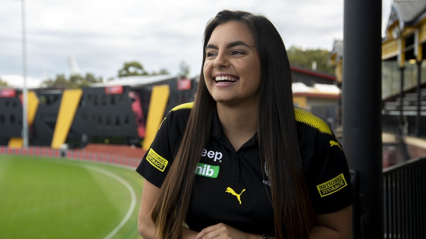 A smiling young woman standing in the grandstand of an AFL football oval.