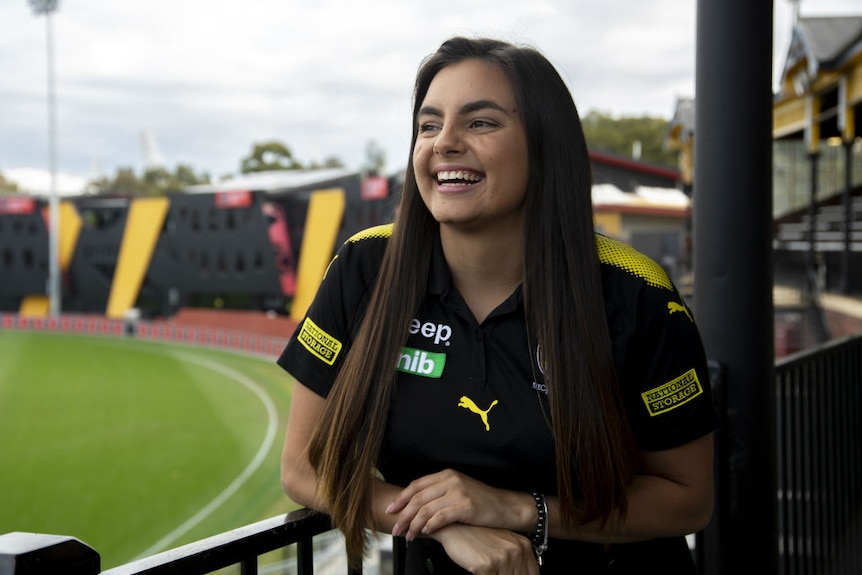A smiling young woman standing in the grandstand of an AFL football oval.