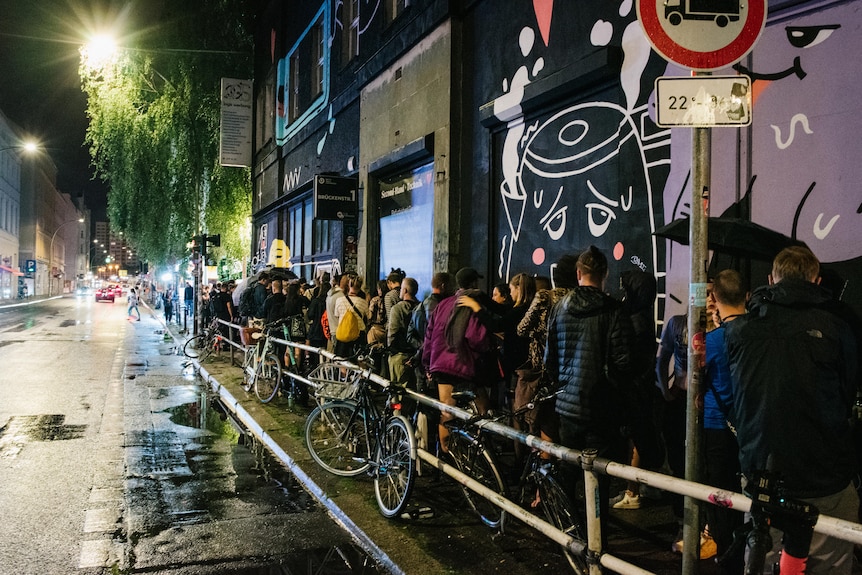 A line of people outside a Berlin nightclub against a grafitti-covered wall.