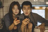 Two people, a man and a woman, in their 20s sit in a cramped space next to a toilet bowl both looking at their smartphones.