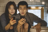 Two people, a man and a woman, in their 20s sit in a cramped space next to a toilet bowl both looking at their smartphones.