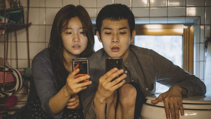 Two people, a man and a woman, in their 20s sit in a cramped space next to a toilet bowl both looking at their smartphones.