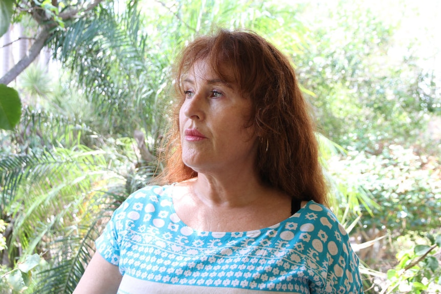 Woman sitting with greenery in the background, she is looking off in the distance and doesn't look happy.