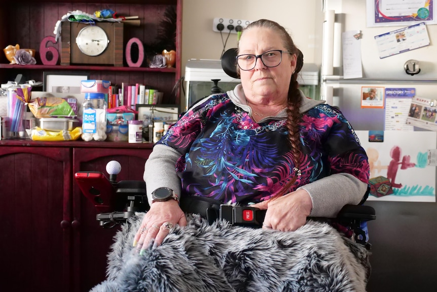 A middle-aged woman sits in her electric wheelchair inside her dining room. 