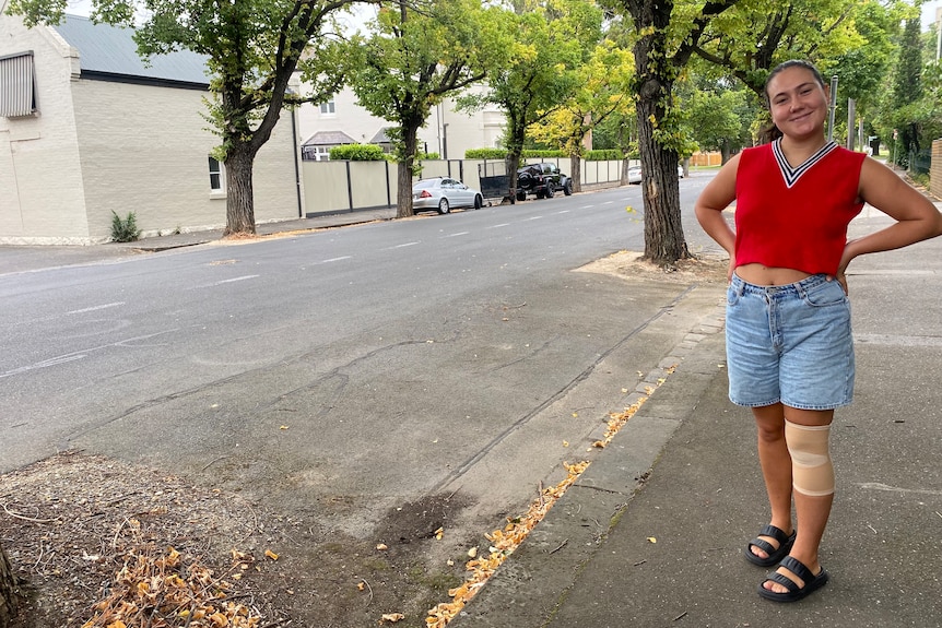 Ella Taverner wears a red singlet and jean shorts while standing, hands on hips, on a footpath and smiling. 