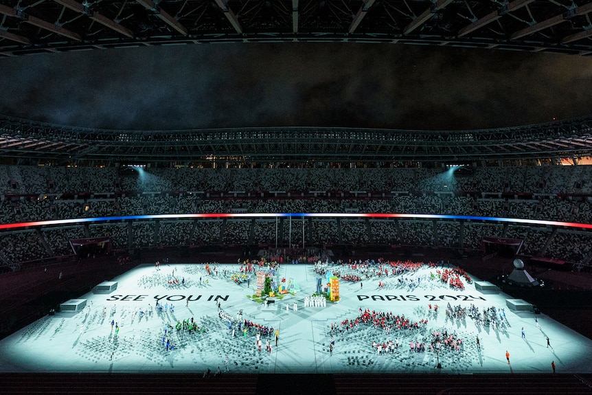 Athletes gather on the illuminated ground of a stadium at night, where the message 'See you in Paris 2024' is projected..