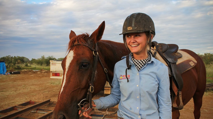 Jaiden Hill and her horse, smiling in front of a stormy sky. Jaiden is dressed in a button-up shirt and sparkly belt.