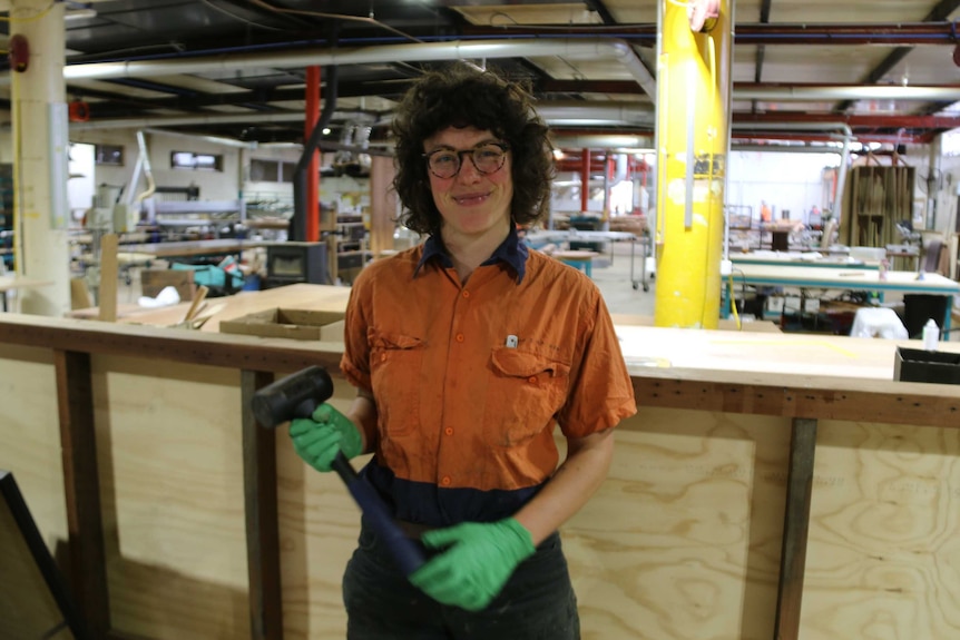 A young woman inside a workshop holds a hammer and stands smiling at the camera.