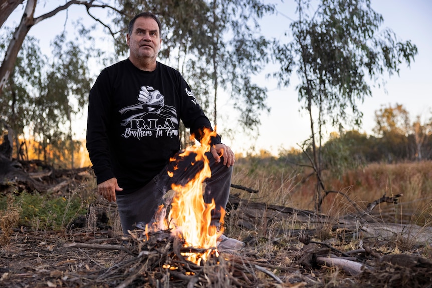 a man kneels before a small fire with dropping eucalyptus branches in the background, 