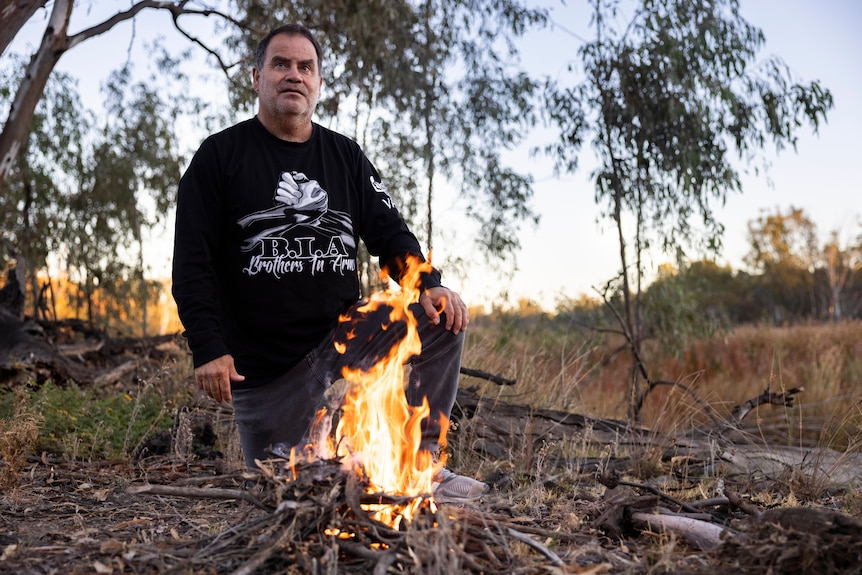 a man kneels before a small fire with drooping eucalyptus branches in the background, 