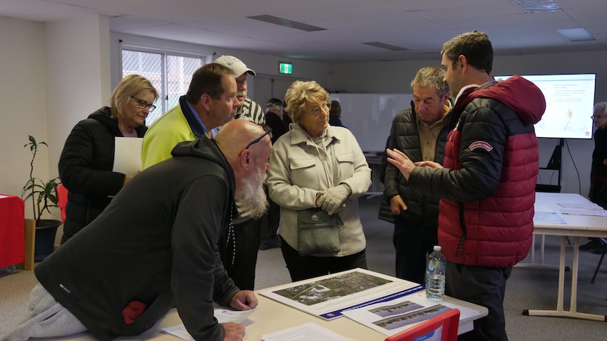 Six annoyed looking people cluster around another man who is explaining documents on a table between them