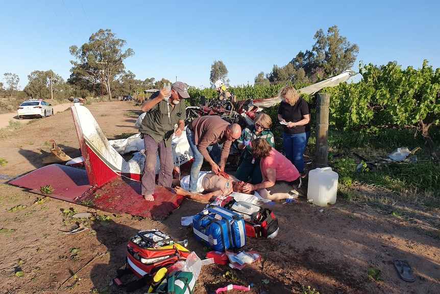 Several people treating an injured man lying on the ground next to a damaged light plane in a vineyard