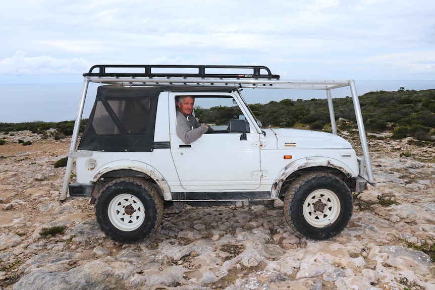 Man looking out window of small white Suzuki with roof racks on a rocky area.