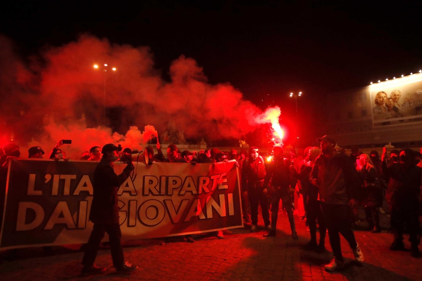 People light flare, while others hold a large sign as they protest against the government restrictions.