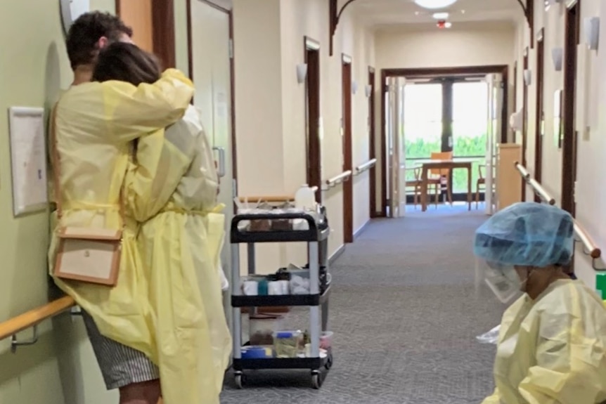 A woman dressed in PPE is comforted by a man, while another woman sits close by