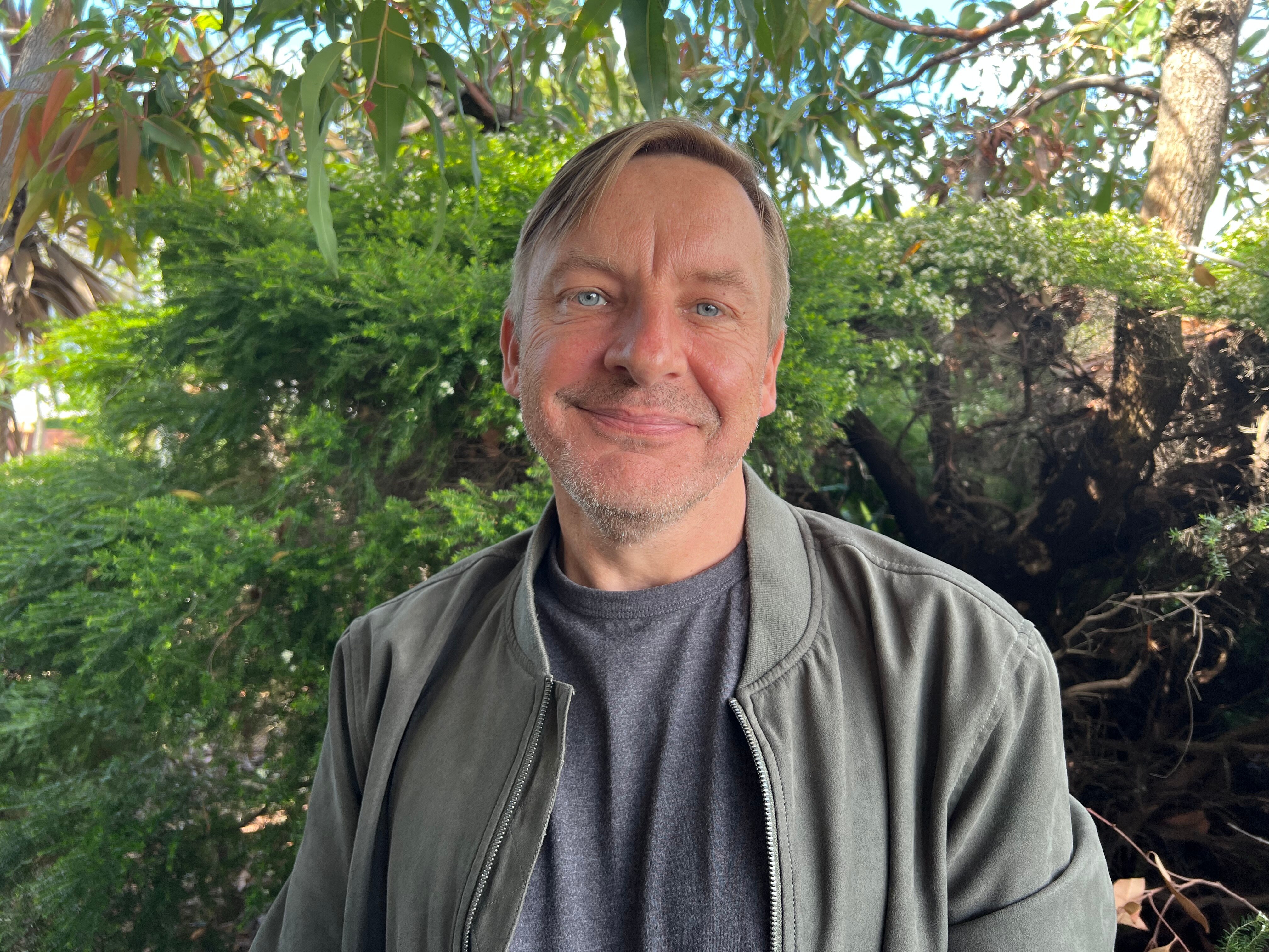 A man with fair hair, wearing black jacket and light shirt, smiles with mouth closed. He's surrounded by bushes.
