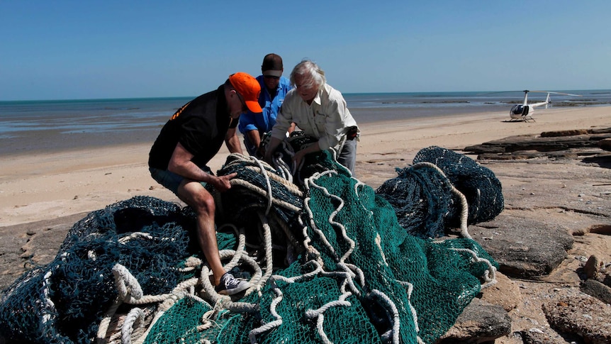 Three men try to lift large green heavy commercial fishing net on rocky part of a beach with clear blue sky above.