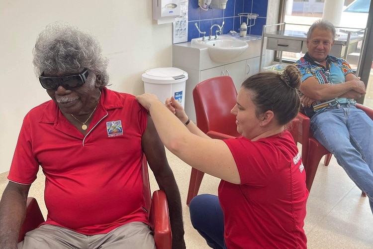 An Aboriginal man sits on a chair in a clinic and receives a COVID vaccine from the nurse knelt beside him.